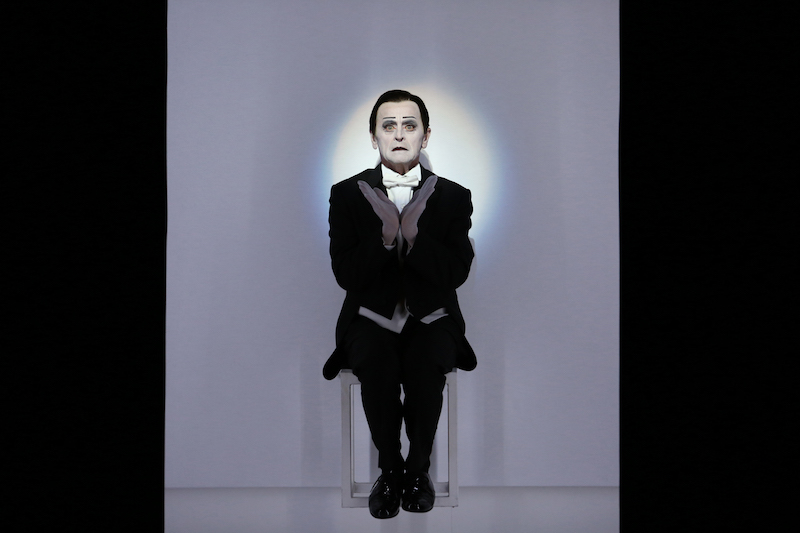 Mikhail Baryshnikov in dramatic stage makeup and tuxedo sits on a white cube. His face is illuminated by a light and the heels of his hands form a V in front of his chest.